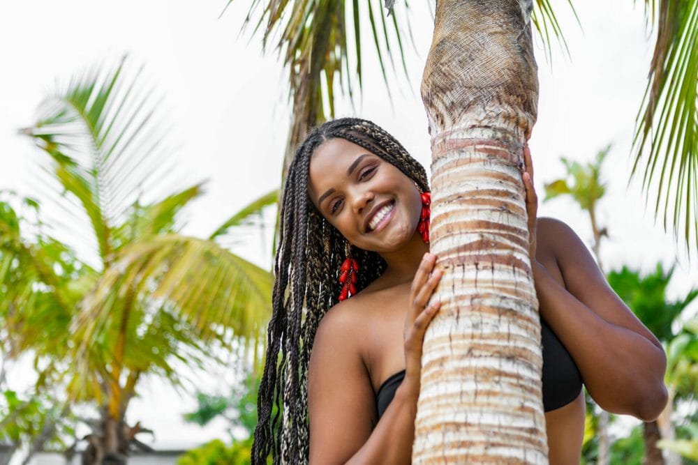 woman standing outdoors behind a tree and smiling with knotless braids
